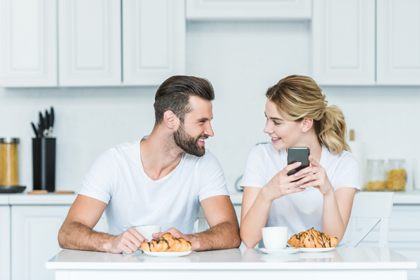 happy young couple smiling each other and using smartphone while having breakfast together - Фото, зображення