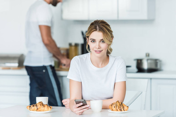 beautiful young woman using smartphone and smiling at camera while boyfriend preparing breakfast behind - Fotografie, Obrázek