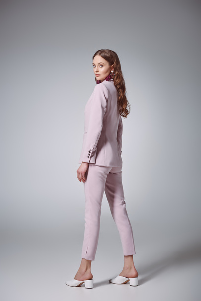 side view of attractive woman in fashionable pink suit looking at camera on grey - Фото, зображення