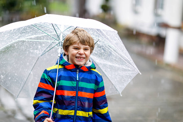 Schöne kleine Junge auf dem Weg zur Schule zu Fuß bei Schneeregen, Regen und Schnee mit einem Regenschirm an kalten Tag. Glückliches und fröhliches Kind in farbenfroher Freizeitkleidung. - Foto, Bild