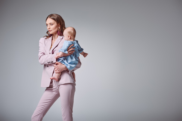 schöne stylische junge Mutter trägt entzückende kleine Tochter und schaut isoliert auf grau - Foto, Bild