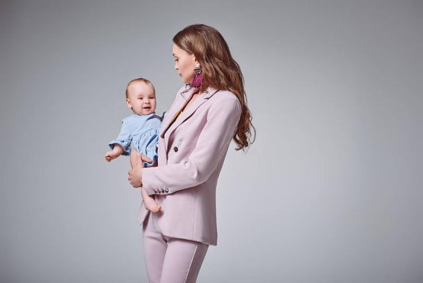 Widok z boku z młoda matka w różowy stylowy garnitur przenoszenia urocza córka niemowlę na szarym tle - Zdjęcie, obraz