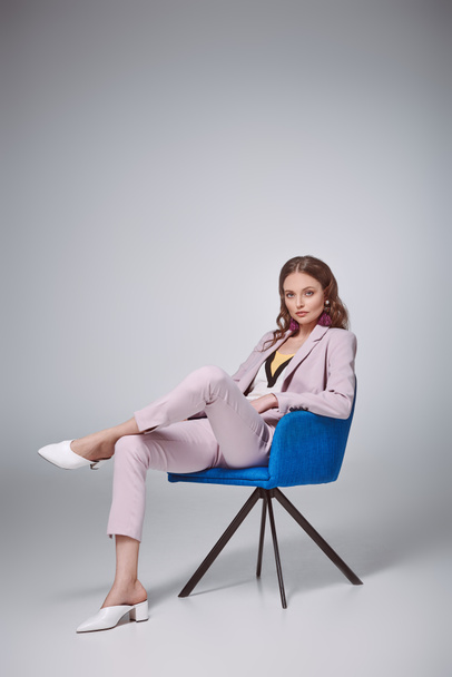 confident stylish woman sitting on blue chair and looking at camera on grey - 写真・画像