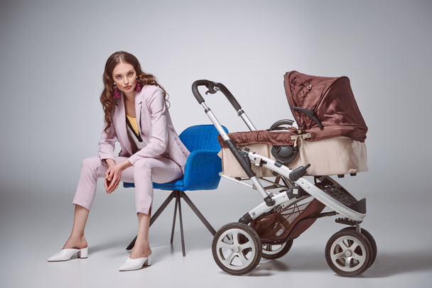 beautiful woman in fashionable suit sitting on chair near baby carriage and looking at camera on grey - Photo, Image