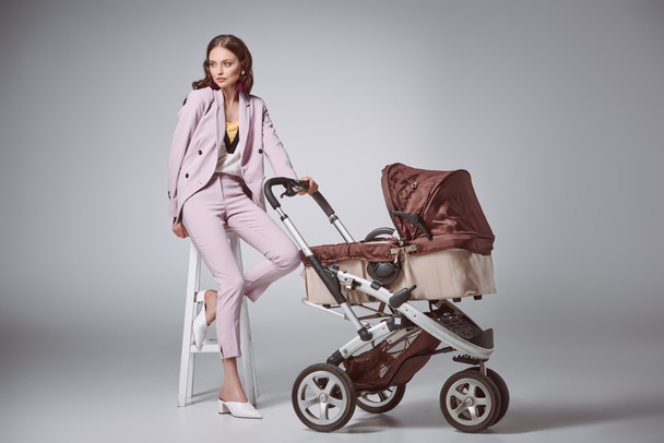 fashionable young woman with baby carriage sitting on stool and looking away on grey - Foto, Imagen