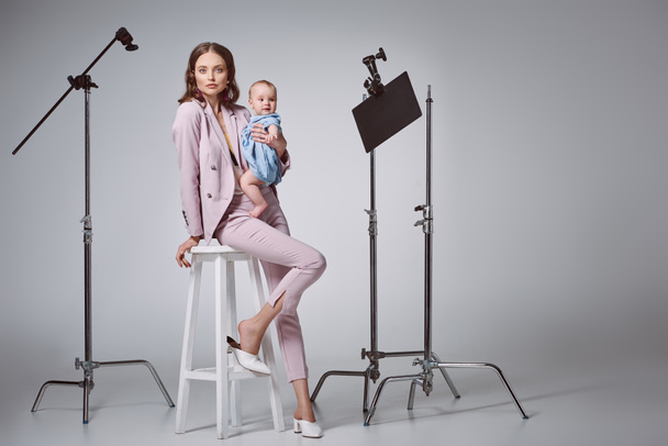 beautiful stylish mother holding adorable infant daughter and looking at camera while sitting on stool in recording studio - Foto, afbeelding