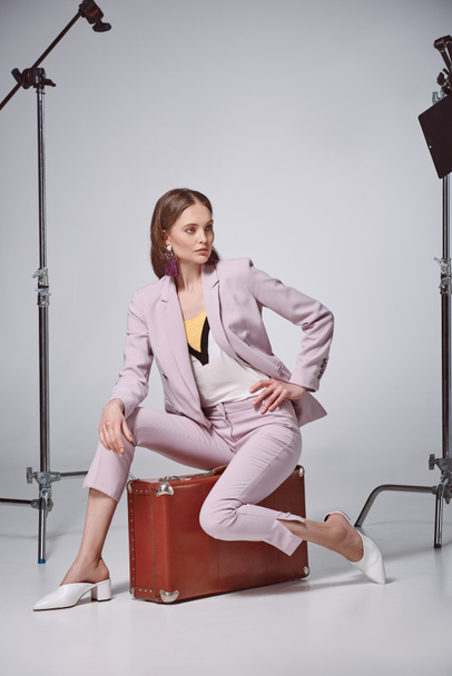 fashionable woman in pink suit sitting on suitcase and looking away in recording studio  - Fotó, kép