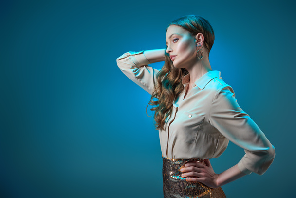 beautiful woman in fashionable golden glittering skirt posing with hand on waist and looking away isolated on blue   - Foto, afbeelding