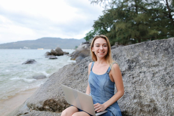 Молодая красивая женщина с ноутбуком, сидящая на камне у моря, пустой утренний пляж
. - Фото, изображение