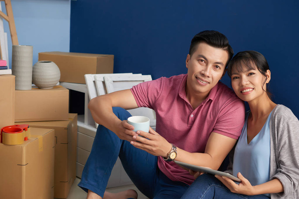 Aasialainen pari hymyilee kameralle onnellisesti istuessaan uudessa asunnossa pakatuissa pahvilaatikoissa
  - Valokuva, kuva