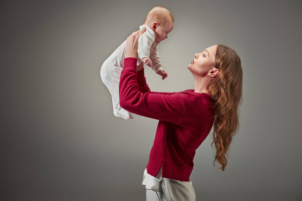 Seitenansicht der schönen jungen Mutter hält entzückende Baby-Mädchen isoliert auf grau - Foto, Bild