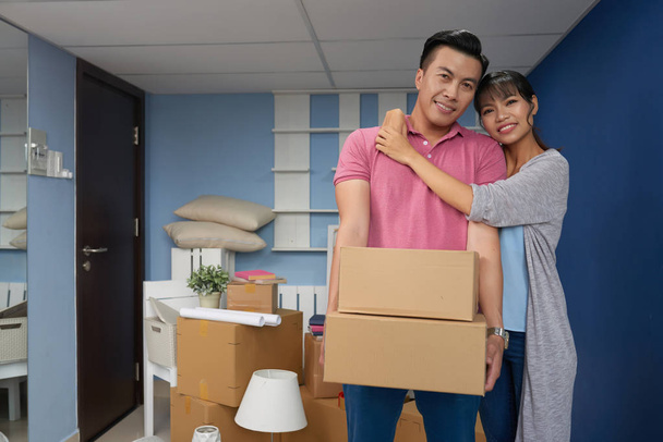 Aasialainen pari seisoo uudessa kodissa, pitäen pahvilaatikoita, joissa on kotitaloustavaroita huoneessa
  - Valokuva, kuva