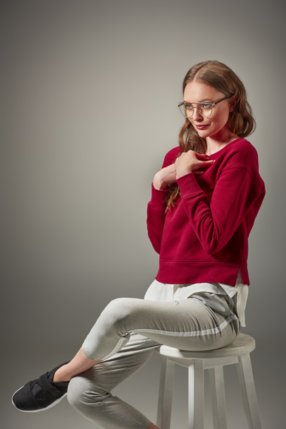 beautiful smiling woman in spectacles sitting on stool and looking away isolated on grey - 写真・画像