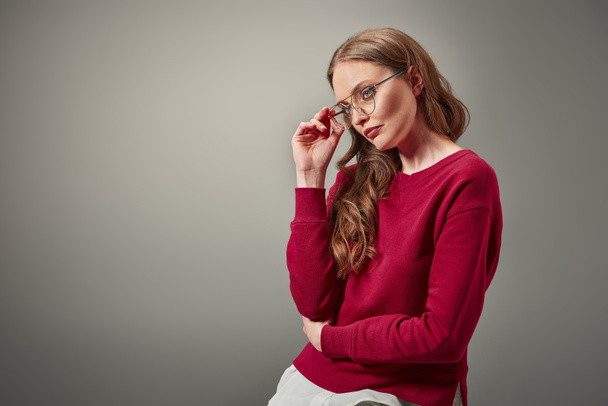 stylish woman adjusting eyeglasses and looking away isolated on grey - Foto, Imagen