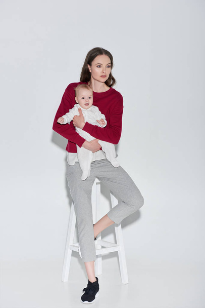 full length view of beautiful stylish woman holding adorable baby girl and sitting stool on grey - Фото, изображение