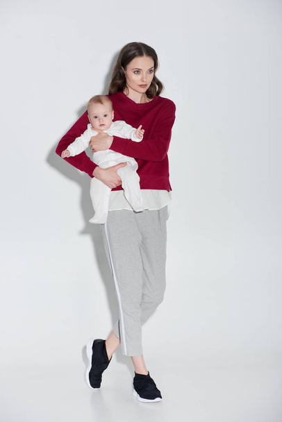 full length view of beautiful young mother in stylish outfit carrying adorable baby girl on grey - Photo, Image