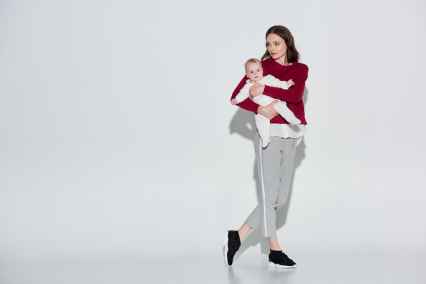full length view of beautiful young mother in stylish outfit carrying adorable infant daughter on grey - Photo, Image
