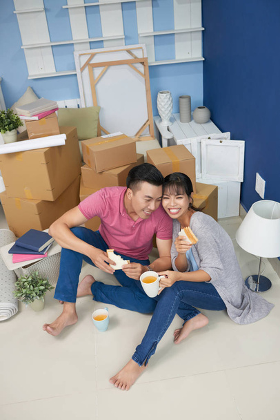 Joven pareja asiática comiendo sándwiches y sonriendo felizmente mientras se sienta en el suelo en una habitación desordenada con cajas de cartón sin embalar
 - Foto, imagen