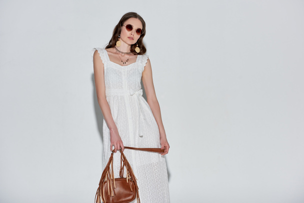 beautiful woman in stylish white dress and sunglasses holding handbag and looking at camera on grey - Photo, Image