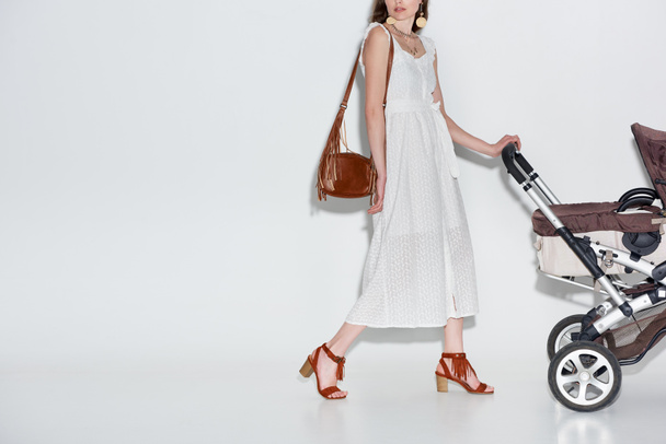 plan recadré de jeune femme en robe blanche à la mode marchant avec chariot bébé sur gris
 - Photo, image