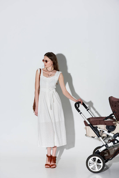 stylish woman in white dress and sunglasses standing with baby carriage and looking away on grey  - Fotó, kép