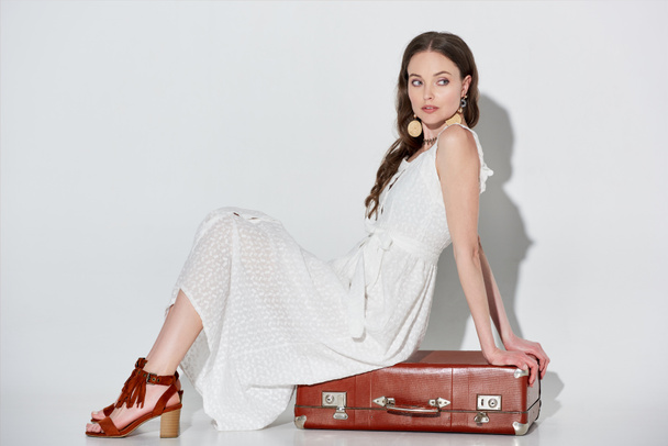 beautiful woman in stylish white dress sitting on retro suitcase and looking away on grey  - Photo, Image