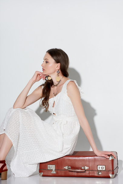 beautiful pensive young woman in stylish white dress sitting on retro suitcase and looking away on grey - Zdjęcie, obraz
