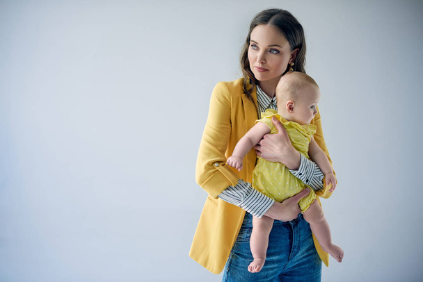 young mother in stylish outfit carrying adorable infant daughter isolated on grey  - Фото, зображення