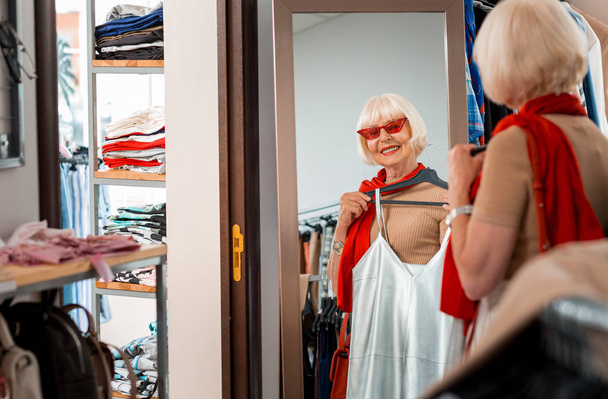 Pleased grey haired woman keeping summer dress in front of her in fitting room of shopping store - Foto, Imagem