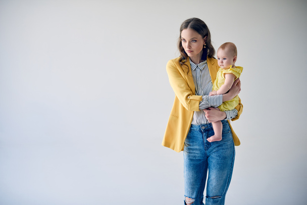 madre joven de moda llevando a su adorable hija lactante y mirando hacia otro lado aislado en gris
 - Foto, imagen
