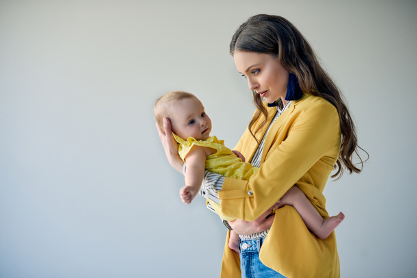 Seitenansicht der schönen stilvollen Mutter trägt entzückende Säugling Tochter isoliert auf grau - Foto, Bild