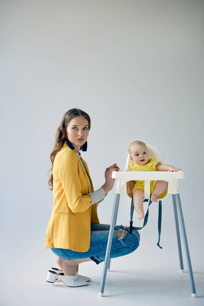 bela mãe na moda agachamento perto da filha infantil sentada em cadeira alta no cinza
  - Foto, Imagem
