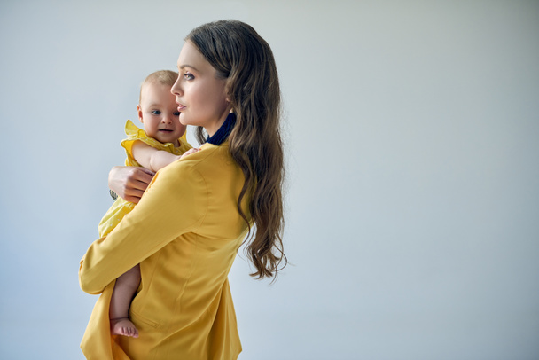 side view of stylish woman carrying adorable baby girl isolated on grey - Photo, Image