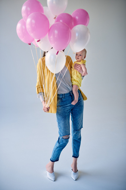 full length view of stylish woman carrying adorable baby girl and holding balloons on grey - Фото, изображение