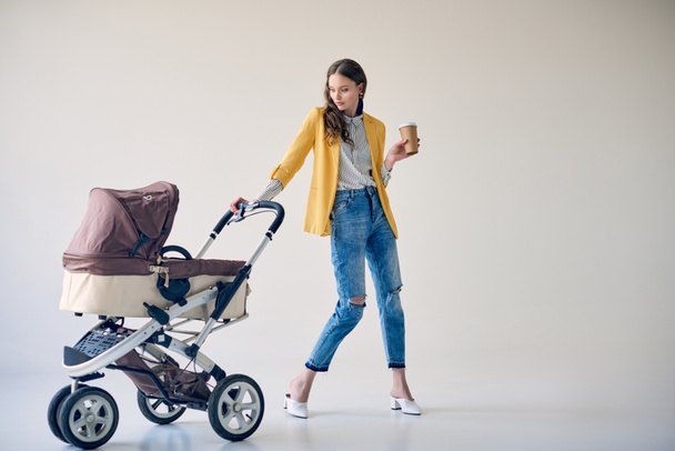 stylish young woman holding disposable coffee cup and looking at baby stroller on grey   - Фото, зображення