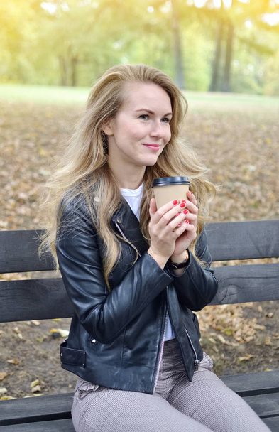 秋の公園で歩いているかなり若い女性リラックス レジャー黒革ジャケット ファッション現代赤い爪飲みコーヒーを離れて - 写真・画像
