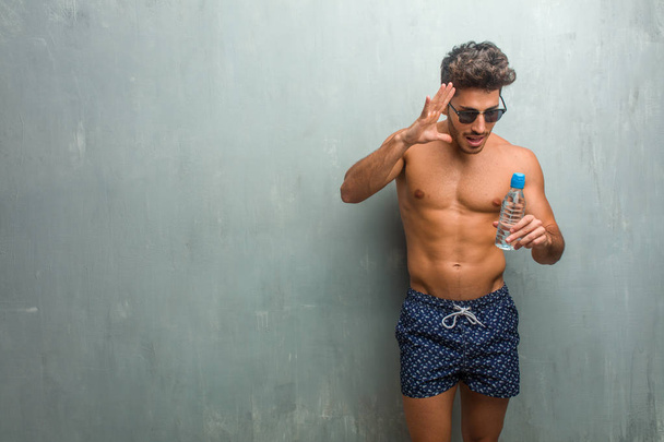 Young athletic man wearing a swimsuit against a grunge wall surprised and shocked, looking with wide eyes, excited by an offer or by a new job, win concept - Foto, Imagen