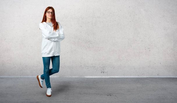 Young redhead girl in an urban white sweatshirt with glasses keeping the arms crossed in frontal position. Confident expression and standing on a vintage gray wall - Photo, Image