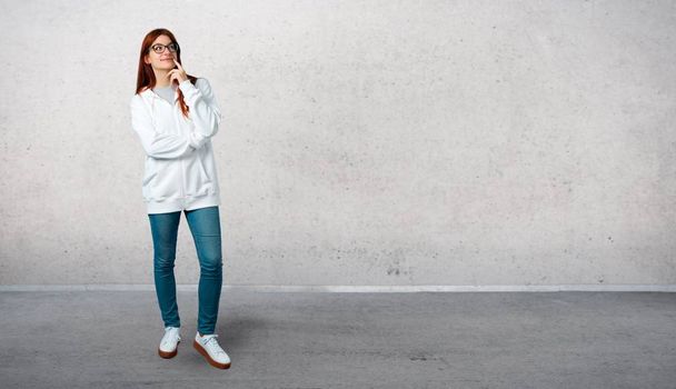 Young redhead girl in an urban white sweatshirt with glasses standing and thinking an idea while looking up and standing on a vintage gray wall - Fotó, kép