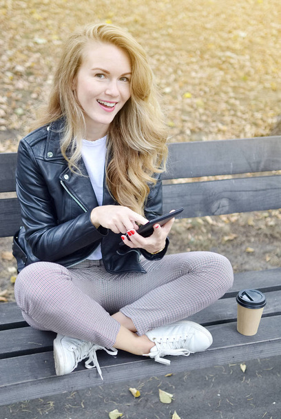 かなり若い女性座っているベンチ秋の公園デジタル デバイスで本を読んでリラックス レジャー黒革ジャケット ファッション現代赤い爪飲みコーヒーを離れて - 写真・画像