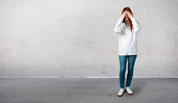 Young redhead girl in an urban white sweatshirt with glasses covering eyes by hands. Do not want to see something and standing on a vintage gray wall - Photo, Image