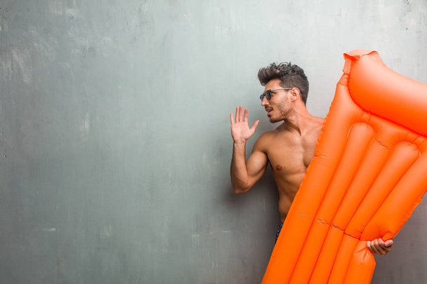 Young athletic man wearing a swimsuit against a grunge wall screaming angry, expression of madness and mental instability, open mouth and half-opened eyes, madness concept - Photo, Image