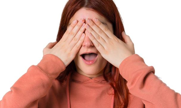 Young redhead girl with pink sweatshirt covering eyes by hands. Surprised to see what is ahead on isolated white background - Φωτογραφία, εικόνα