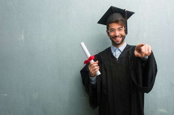 Young graduated man against a grunge wall with a copy space cheerful and smiling pointing to the front - Foto, Bild