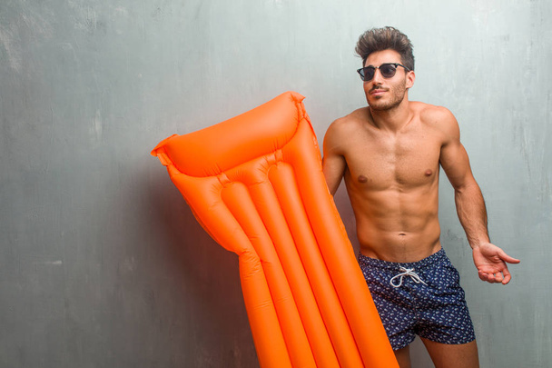 junger athletischer Mann im Badeanzug vor einer Grunge-Wand, zweifelnd und achselzuckend, Konzept der Unentschlossenheit und Unsicherheit, unsicher über etwas - Foto, Bild
