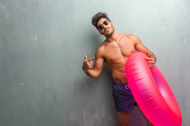 Young athletic man wearing a swimsuit against a grunge wall proud and confident, pointing fingers, example to follow, concept of satisfaction, arrogance and health - Foto, imagen