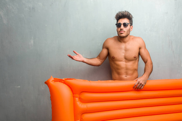 Young athletic man wearing a swimsuit against a grunge wall doubting and shrugging shoulders, concept of indecision and insecurity, uncertain about something - Photo, Image