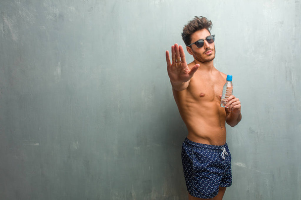 Молодой спортсмен в купальнике против гранж-стены серьезный и решительный, положив руку впереди, остановить жест, отрицать концепцию
 - Фото, изображение