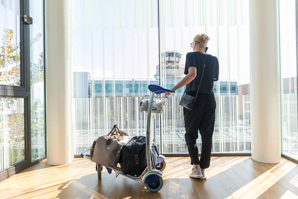 Случайная женщина на терминале аэропорта разговаривает по мобильному телефону, ждет вылета рейса с багажом на тележке
. - Фото, изображение