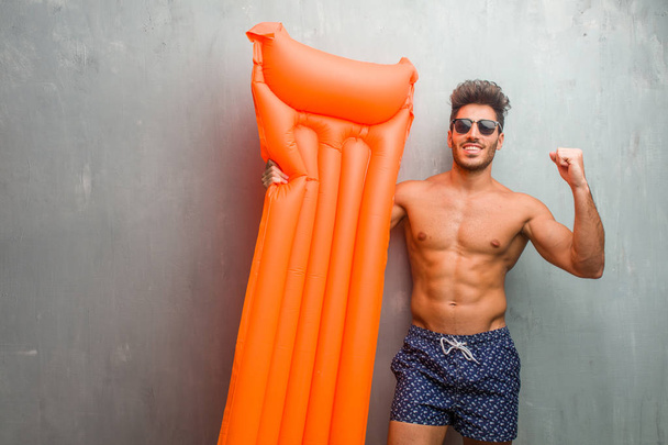 junger, athletischer Mann im Badeanzug vor einer Grunge-Wand sehr glücklich und aufgeregt, die Arme in die Höhe reckend, einen Sieg oder Erfolg feiernd, Lottogewinn - Foto, Bild